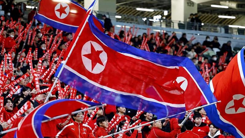 Grupo numeroso de torcedores vestidos de vermelho agita bandeiras da Coreia do Norte em estádio coberto. Muitos seguram bastões infláveis vermelhos enquanto acompanham evento esportivo.