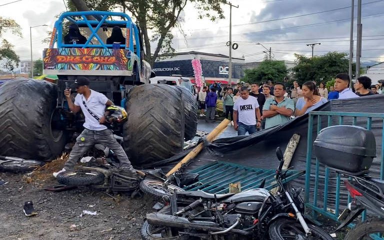 Monster truck azul com pneus gigantes colide com motocicleta preta caída e caminhonete verde danificada. Pessoas observam a cena em rua com árvores e postes ao fundo.