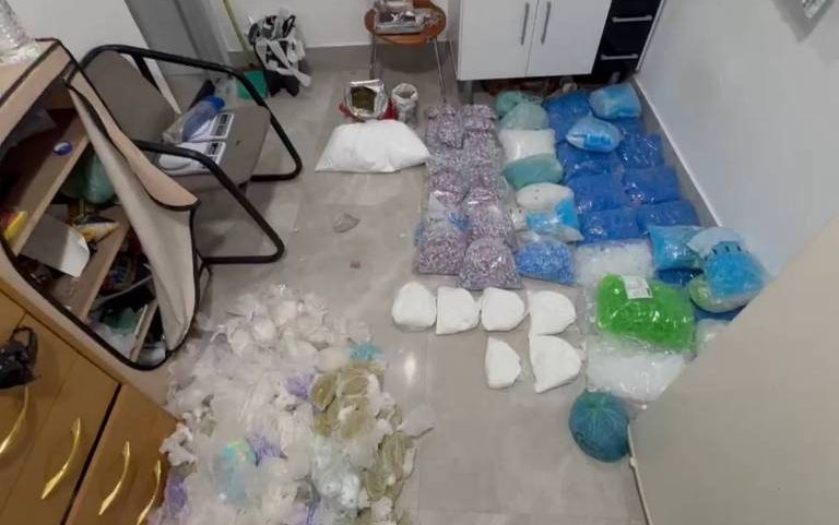Diversos recipientes plásticos, como potes e tampas, estão organizados em fileiras no chão de uma sala. À esquerda, há uma cadeira reclinável e uma cômoda com gavetas. O ambiente tem piso claro e paredes brancas.