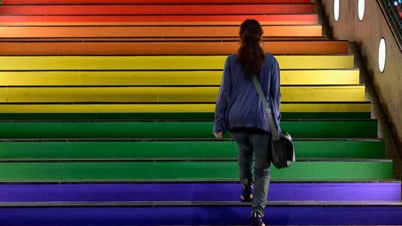 Pessoa de costas, vestindo blusa azul e calça jeans, sobe escada com degraus pintados em cores do arco-íris, do roxo ao vermelho, em ambiente interno com iluminação circular nas paredes laterais.