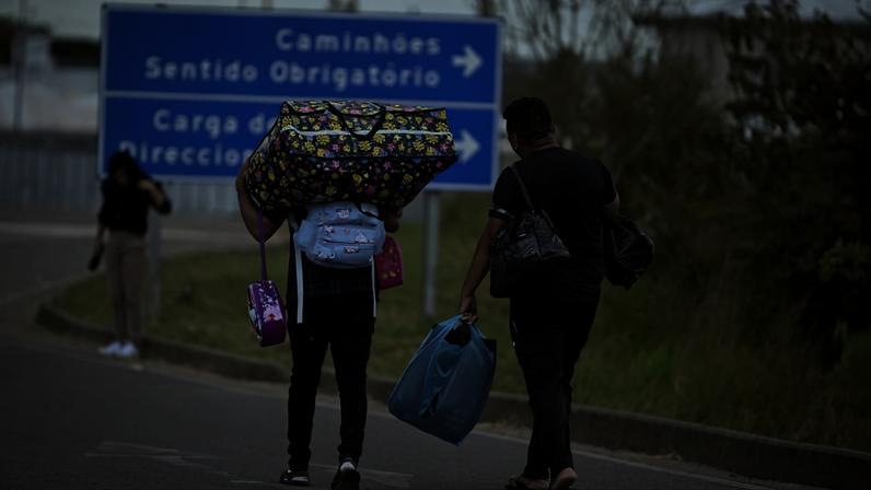 Duas pessoas caminham em estrada carregando grandes malas coloridas na cabeça e bolsas nas mãos. Ao fundo, uma placa azul indica direções para caminhões e carga. Outra pessoa está parada na calçada, usando máscara.