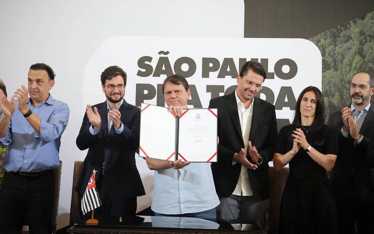 Homem no centro segura documento assinado aberto, cercado por seis pessoas aplaudindo. Ao fundo, painel com texto 'São Paulo pra Toda'. Bandeira de São Paulo está sobre a mesa à frente.