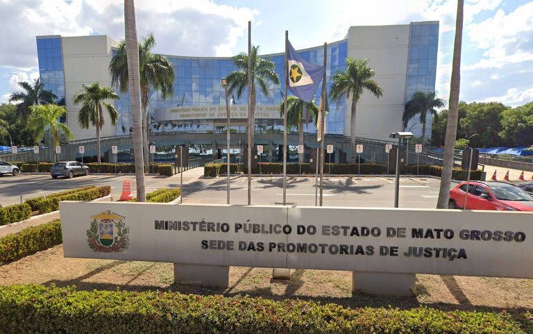 Fachada moderna do prédio do Ministério Público do Estado de Mato Grosso com vidro refletivo e palmeiras na frente. Placa em primeiro plano indica a sede das Promotorias de Justiça. Bandeira do estado hasteada ao centro.