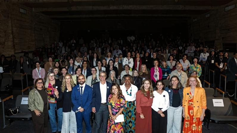 Mais de cem pessoas posam para foto em auditório com iluminação amarela nas paredes. Grupo diversificado está distribuído em fileiras, com dez pessoas em destaque na frente, algumas vestindo roupas formais e outras coloridas.