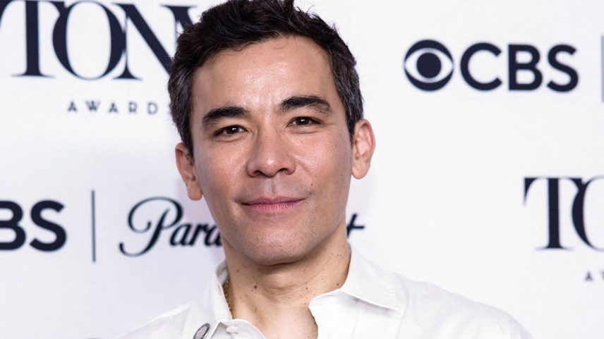 Homem com cabelo curto e escuro veste camisa branca aberta na altura do peito, posando em frente a painel com logos do Tony Awards, CBS e Paramount+. Fundo branco com texto preto.