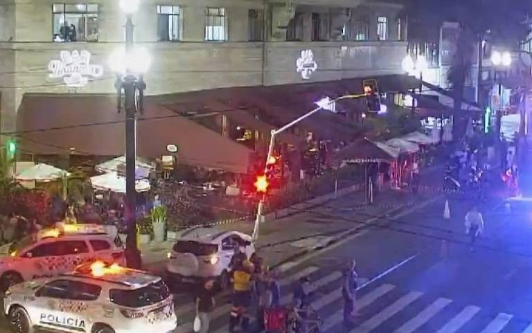 Viaturas policiais com luzes de emergência acesas bloqueiam cruzamento movimentado entre avenidas Ipiranga e São João à noite. Pessoas e veículos estão parados, e prédios iluminados cercam a área.