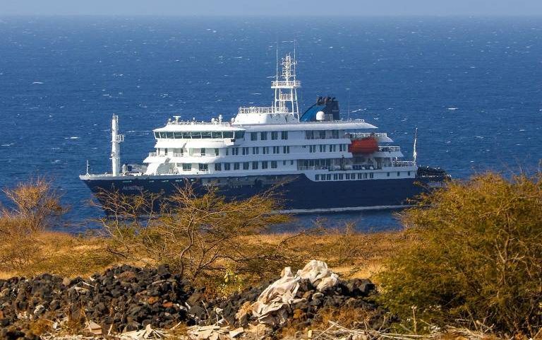 O navio de cruzeiro branco e azul navegando próximo a uma costa com vegetação seca, arbustos e pedras escuras. O mar está calmo e azul ao fundo.