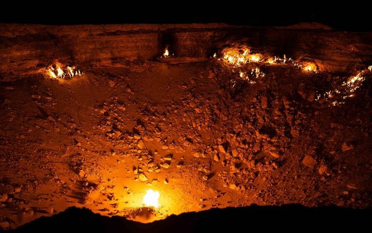 Poço profundo no deserto com chamas ardendo em vários pontos ao longo da borda e no fundo, iluminando a área rochosa ao redor durante a noite.