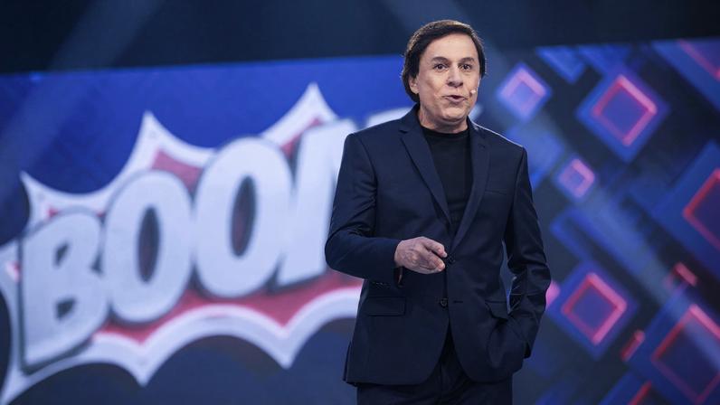 Homem vestido com terno escuro e camiseta preta está em pé em estúdio com fundo azul e a palavra 'BOOM' em destaque atrás dele.