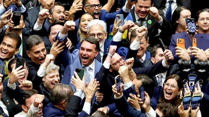 Homens e mulheres em trajes formais comemoram com expressões de alegria e punhos erguidos, muitos segurando celulares para registrar o momento em ambiente interno.