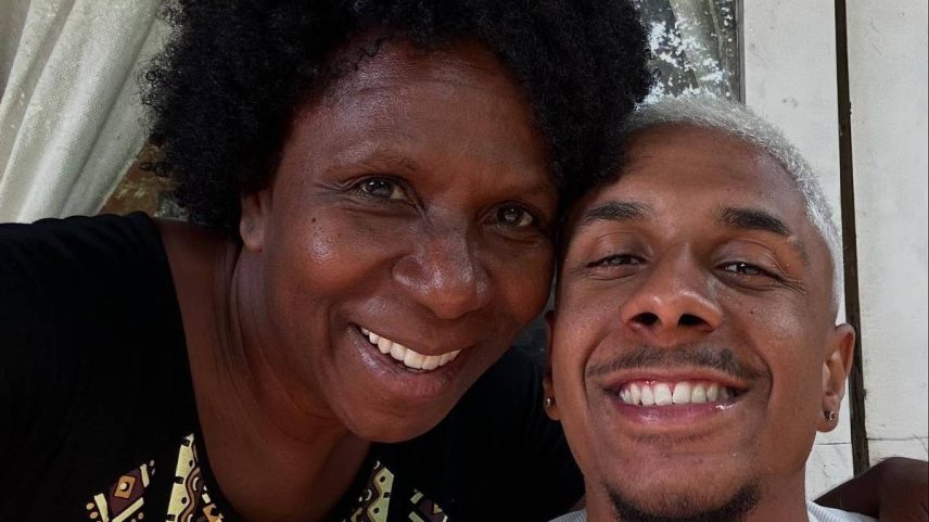 Duas pessoas posam próximas, sorrindo para a câmera. A pessoa à esquerda tem cabelo curto e cacheado, veste blusa preta com detalhes coloridos. A pessoa à direita tem cabelo curto e claro, usa regata branca. Ao fundo, cortinas claras e porta de vidro.