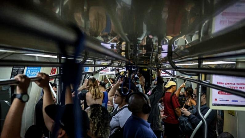 Interior de vagão de metrô cheio de passageiros em pé segurando barras e alças. Pessoas usam roupas casuais e alguns usam fones de ouvido. Luz artificial ilumina o ambiente fechado.