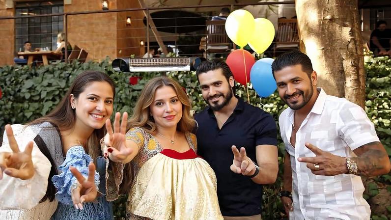 Cinco pessoas posam juntas em ambiente externo com decoração de festa, incluindo balões amarelos, vermelhos e azuis ao fundo. Quatro delas fazem sinal de paz com as mãos. O homem ao centro veste camisa preta e calça bege, e está ao lado de uma árvore e de uma parede com plantas.