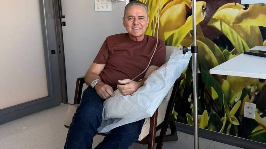 Homem sorridente sentado em cadeira reclinável com soro intravenoso conectado ao braço direito. Ao fundo, parede decorada com pintura de folhas amarelas e mesa hospitalar ao lado.