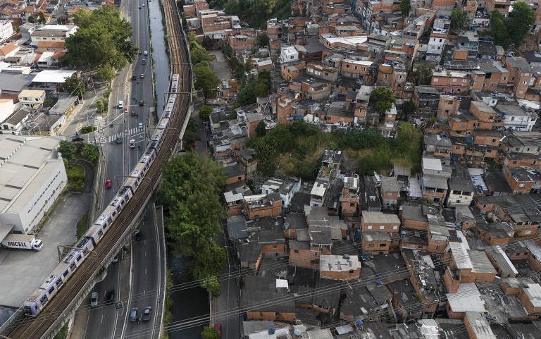 Vista aérea mostra favela com casas pequenas e amontoadas ao lado de linha de trem elevada. Trem branco circula na via férrea paralela a uma avenida com poucos carros. Vegetação dispersa aparece entre as construções e ao longo da linha férrea.