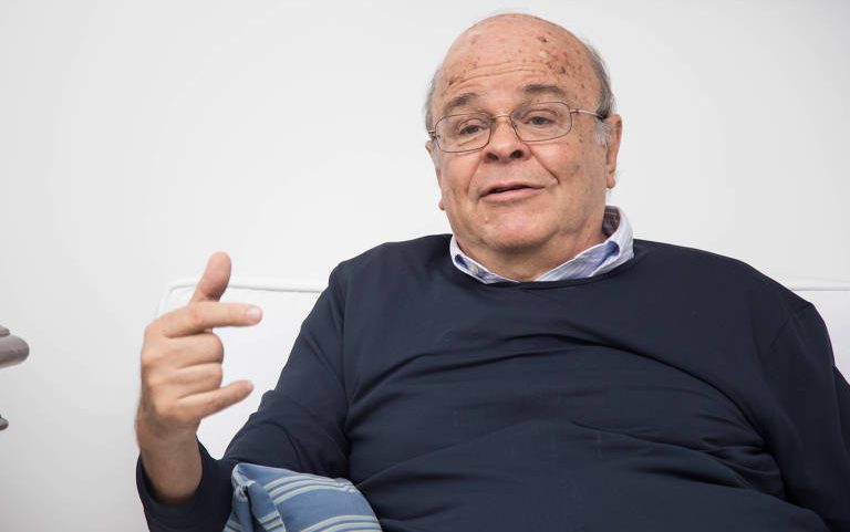 Homem idoso de pele clara, calvo com cabelos ralos nas laterais, usando óculos e camisa azul clara sob suéter azul escuro, sentado em sofá branco, gesticulando com a mão direita levantada. Almofada azul com listras está apoiada em seu braço esquerdo. Fundo branco liso.