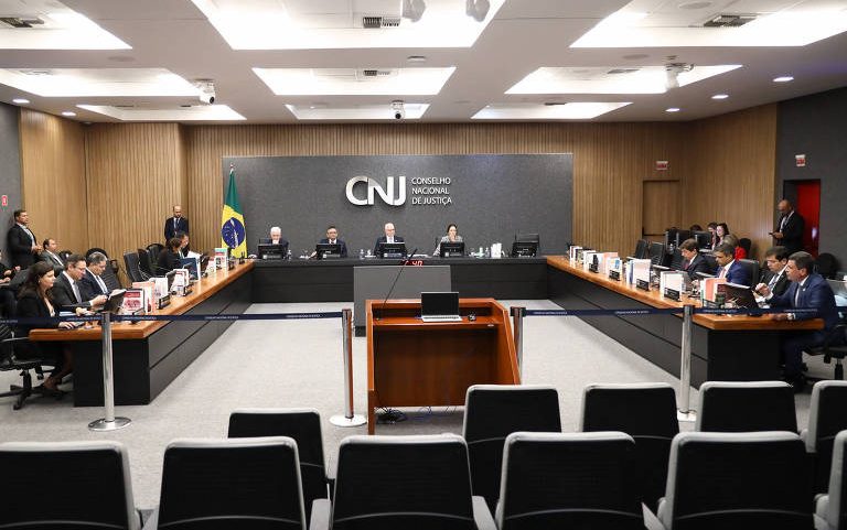 Plenário do Conselho Nacional de Justiça com membros sentados em mesas em formato de U. Bandeira do Brasil ao fundo e painel com logo do CNJ. Ambiente formal com cadeiras vazias em primeiro plano.