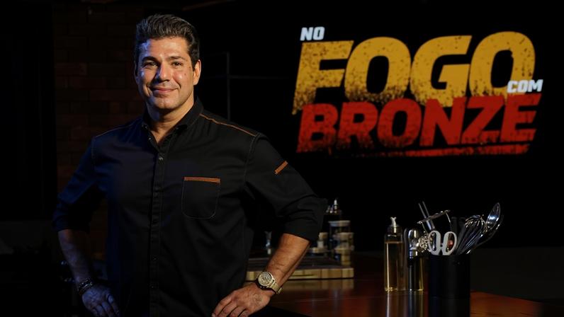 Homem de pé com as mãos na cintura em estúdio de cozinha profissional. Ao fundo, logo iluminado do programa 'No Fogo com Bronze'. Ambiente com iluminação quente e bancada com utensílios de cozinha.