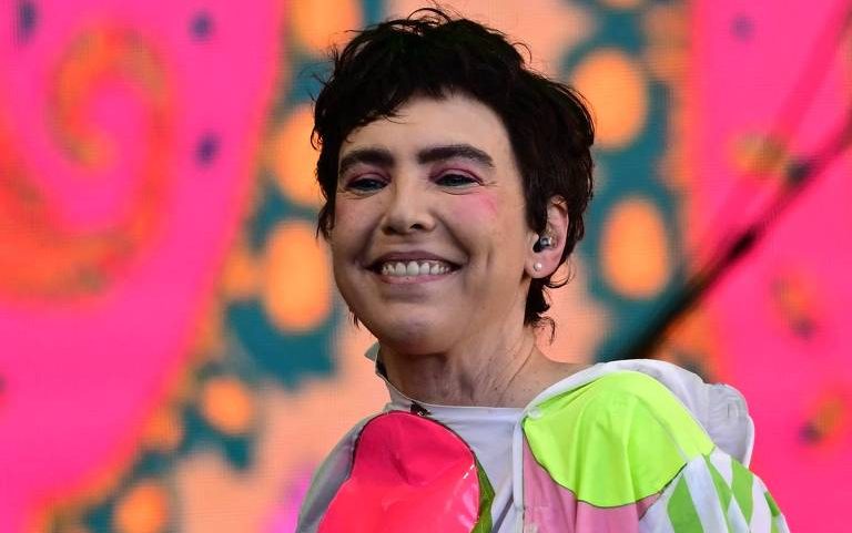 Pessoa com cabelo curto e escuro sorri, usando maquiagem rosa nos olhos e bochechas. Veste roupa com cores neon, incluindo verde e rosa, com fundo desfocado em tons de rosa e laranja.