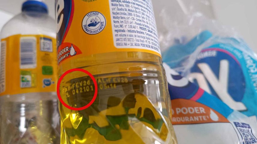 Garrafa plástica de óleo de cozinha com rótulo amarelo exibe data de validade impressa em amarelo na parte transparente do frasco. Ao fundo, outras embalagens plásticas, incluindo uma garrafa e um pacote azul e branco.
