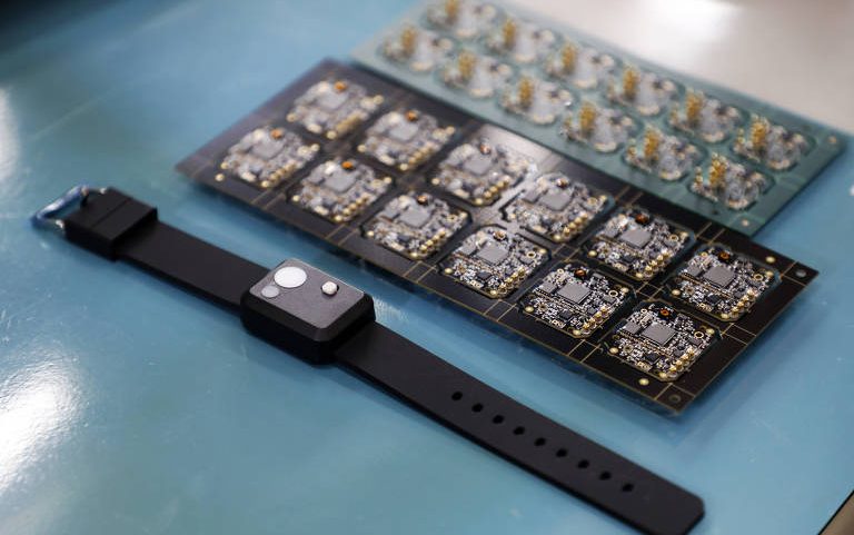Relógio inteligente com pulseira preta e visor quadrado repousa sobre bancada azul ao lado de duas placas de circuito eletrônico com múltiplos componentes montados.