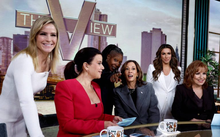 Seis mulheres posam no estúdio do programa The View com a skyline de Nova York ao fundo. Elas estão ao redor de uma mesa com canecas e papéis, vestindo roupas formais em cores variadas.