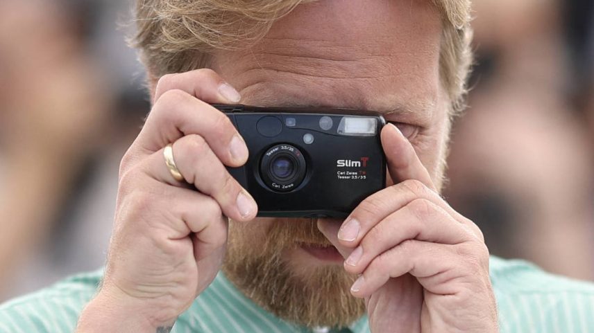Homem de cabelo loiro e barba usa câmera compacta preta para tirar foto. Ele veste camisa verde listrada e relógio preto no pulso esquerdo.