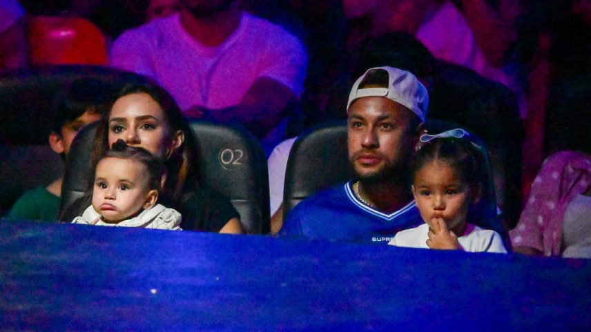 Homem com boné branco para trás e mulher sentados lado a lado em cadeiras numeradas, cada um segurando uma criança pequena. Fundo com outras pessoas em ambiente pouco iluminado. Neymar fecha sessão do circo Stankovic para sua família e amigos em Santos e causa tumulto ao deixar o espetáculo