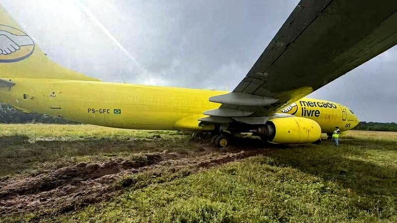 Avião amarelo da Mercado Livre parado em campo de grama com marcas de pneus no solo. Parte da asa e motor visíveis, céu nublado ao fundo.