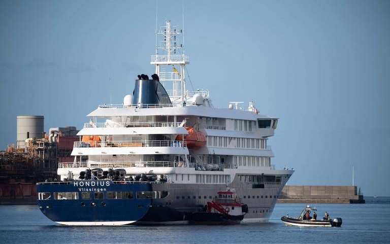 Navio de cruzeiro branco e azul chamado Hondius está atracado em porto com estrutura industrial ao fundo. Duas pequenas embarcações de apoio, uma vermelha e outra preta, estão próximas ao navio na água calma sob céu claro.