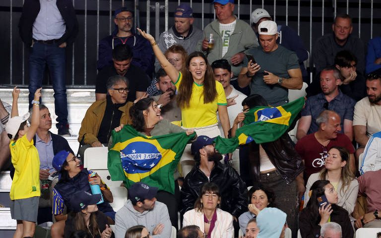 Grupo de torcedores brasileiros em arquibancada, alguns com camisas amarelas e bandeiras do Brasil, celebrando e vibrando. Pessoas ao redor observam e interagem, ambiente noturno com iluminação artificial.