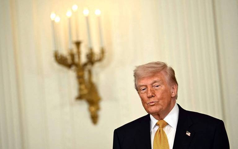 Homem de cabelo loiro e terno escuro com gravata amarela está em ambiente interno com parede clara e candelabro dourado aceso ao fundo.