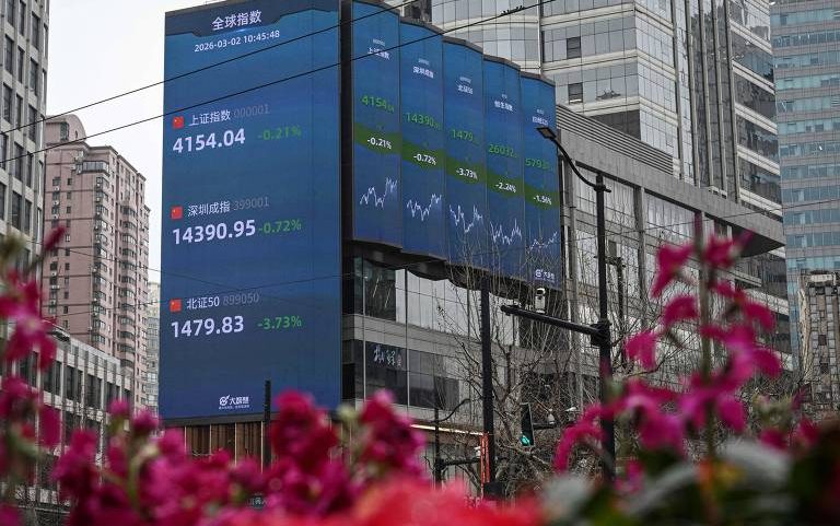 Painel eletrônico grande em fachada de prédio mostra índices financeiros com valores e variações percentuais em verde. Edifícios altos cercam o local, com flores vermelhas desfocadas em primeiro plano.