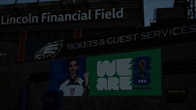 Fachada do Lincoln Financial Field em dia claro, com letreiro grande e banner promocional da Copa do Mundo FIFA 2026. Banner exibe jogador da seleção dos EUA, texto 'WE ARE 26' e troféu da FIFA, com referência a Philadelphia.