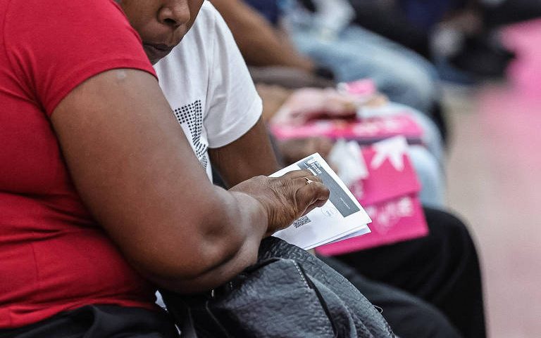 Duas pessoas sentadas lado a lado conferem documentos em papel. Uma veste camiseta vermelha e segura uma bolsa preta, enquanto a outra veste camiseta branca. Ao fundo, outras pessoas também estão sentadas, algumas com pastas ou papéis nas mãos.