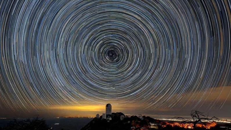 Rastros circulares de estrelas no céu noturno formam um padrão concêntrico acima de um observatório em colina. Luzes da cidade iluminam o horizonte ao fundo.