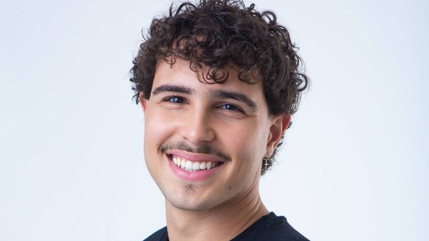 Jovem de cabelos cacheados e curtos sorri para a câmera, vestindo camiseta preta, com fundo branco neutro.