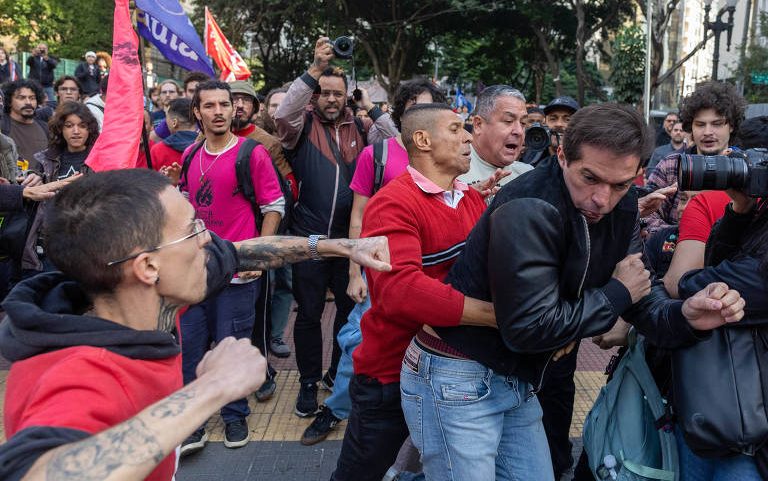 Homem de camiseta vermelha avança com soco em direção a outro homem de jaqueta preta que se protege com os braços. Pessoas ao redor observam e registram a cena com celulares e câmeras. Bandeiras e árvores aparecem ao fundo.