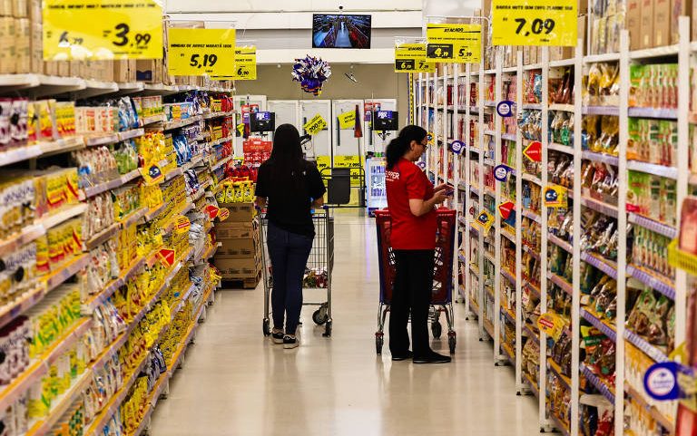 Duas mulheres empurram carrinhos em corredor de supermercado entre prateleiras cheias de produtos alimentícios embalados. Placas amarelas indicam preços em reais.