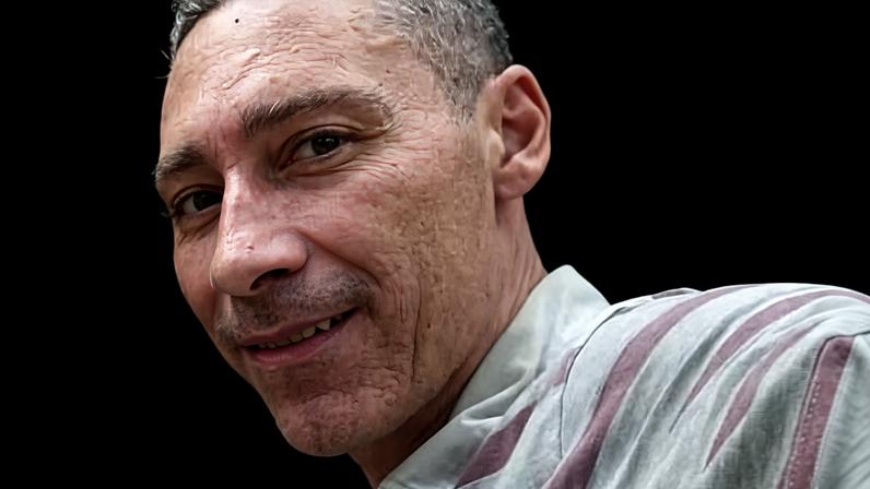 Homem de pele clara e cabelo curto sorri levemente para a câmera. Ele veste camisa polo listrada em tons de cinza e vinho, e está em um ambiente com estrutura metálica e fiação ao fundo.