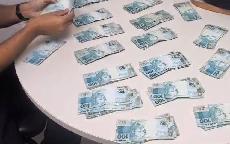 Mãos de uma pessoa organizam várias cédulas de 100 reais espalhadas sobre uma mesa redonda branca. Ao lado, há um objeto preto, possivelmente uma bolsa ou estojo.