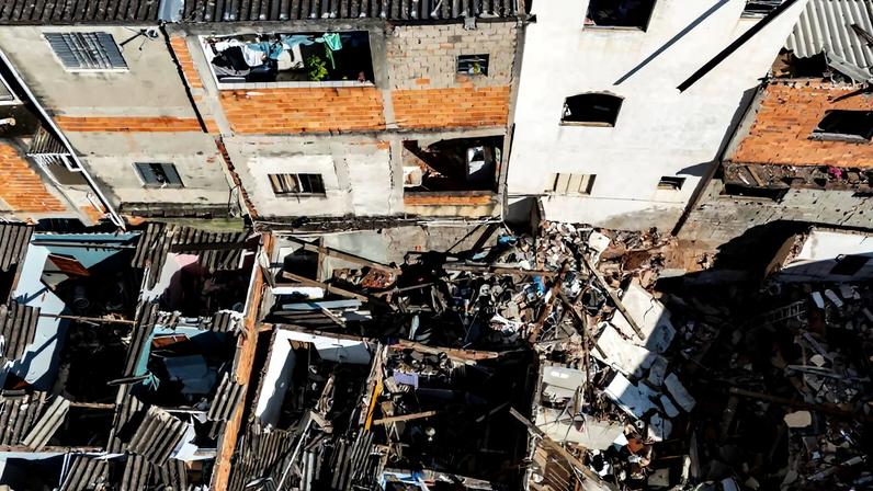 Vista aérea mostra casas parcialmente destruídas com escombros e destroços acumulados. Residências vizinhas permanecem intactas, evidenciando danos concentrados em uma área específica.