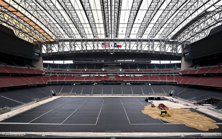 Estádio com teto retrátil aberto parcialmente, arquibancadas vazias e campo coberto por lona preta. Equipamentos e areia estão no lado direito do campo, indicando montagem em andamento.