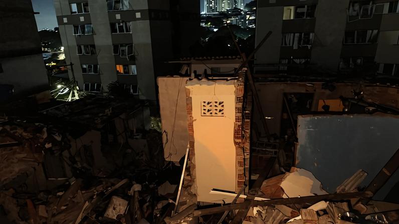 Restos de um edifício residencial demolido à noite, com escombros espalhados e uma parede remanescente iluminada. Prédios altos ao fundo e luzes da cidade visíveis.