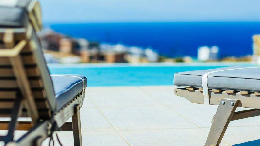 Duas espreguiçadeiras com almofadas cinza estão posicionadas ao redor de uma piscina de borda infinita. Ao fundo, há vista para o mar azul e construções urbanas desfocadas sob céu claro.