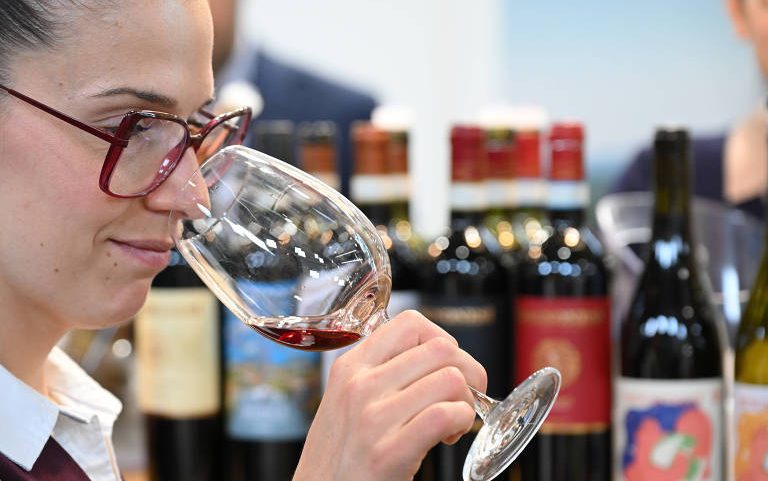 Pessoa com óculos de armação vermelha segura taça com vinho tinto próximo ao nariz, realizando análise olfativa. Ao fundo, várias garrafas de vinho alinhadas em prateleira.