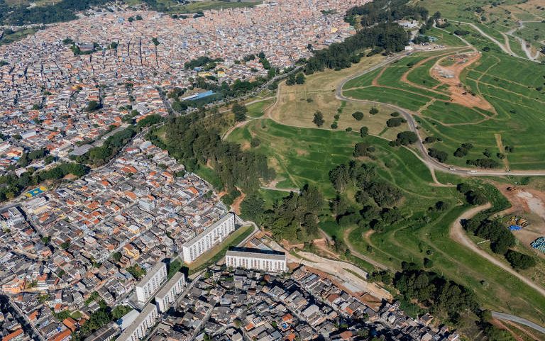 Vista aérea mostra área urbana densa com casas e prédios baixos à esquerda e área de antigo aterro árvores à direita, em São Paulo.