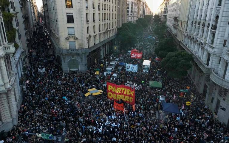 Milhares de pessoas ocupam avenida entre prédios altos em manifestação. Bandeiras vermelhas e faixas com inscrições políticas são visíveis no centro do grupo.