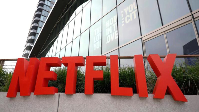 Letreiro grande com a palavra 'NETFLIX' em letras vermelhas maiúsculas fixado em parede clara, com prédio de vidro refletindo o céu ao fundo.