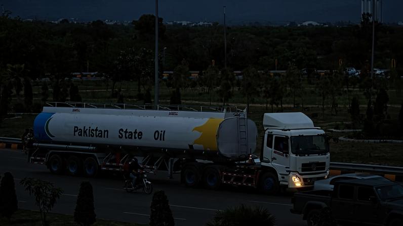Caminhão-tanque branco com inscrição "Pakistan State Oil" trafega por avenida com vegetação e árvores ao fundo. Motociclista e outros veículos circulam próximos.